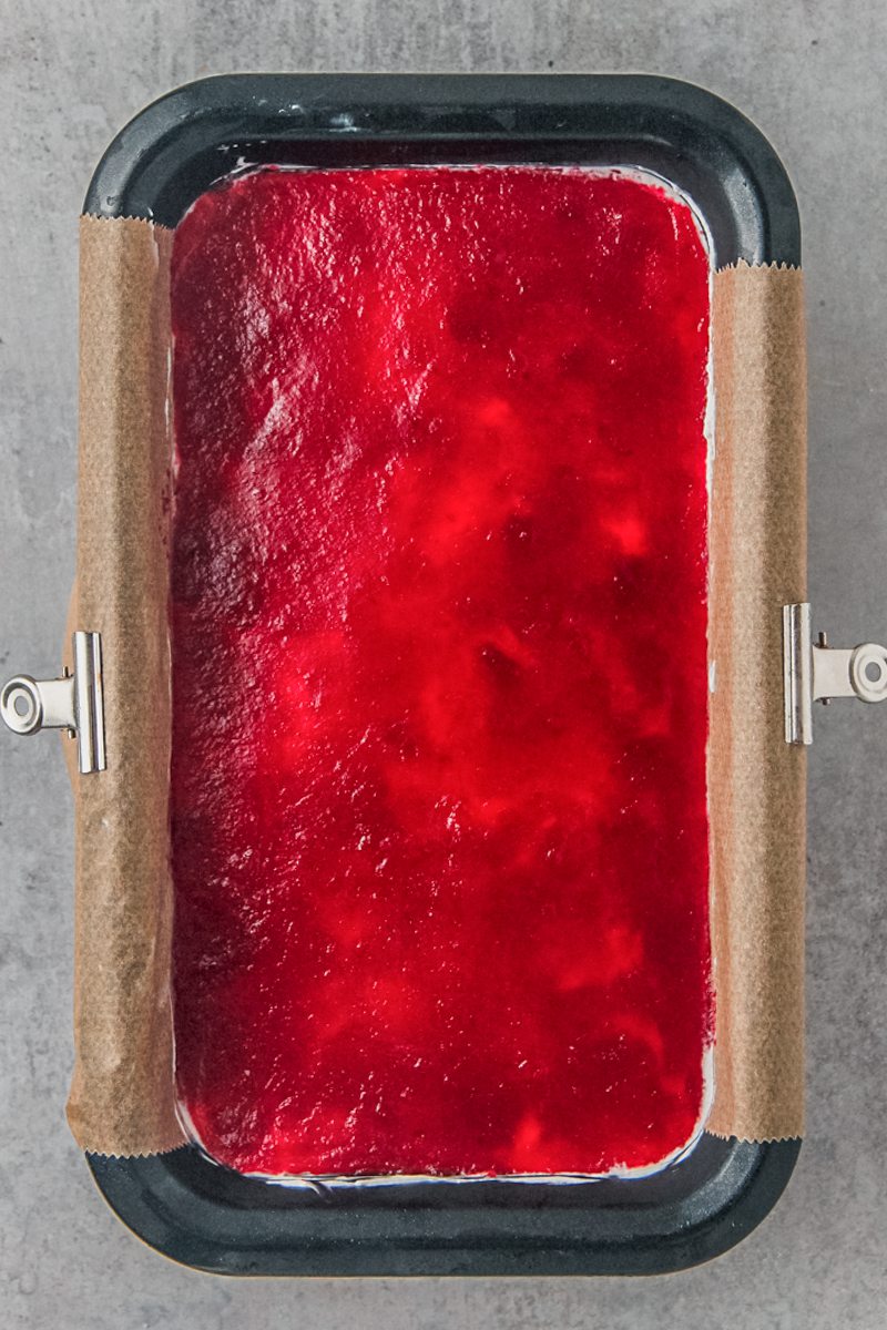 A smooth layer of cranberry puree sits on top of a cranberry cheesecake in a metal loaf tin on a gray surface.