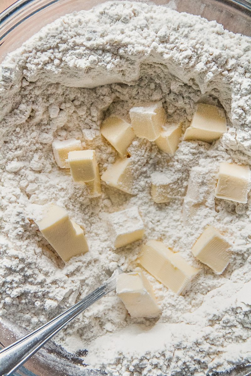 Cubes of butter sits in a well of dry ingredients in a glass bowl on a wooden surface.