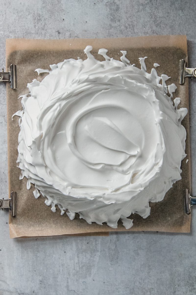 An unbaked pavlova sits on a baking tray lined with greaseproof paper on a gray surface.