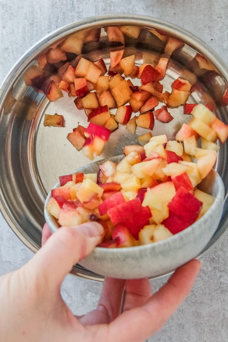 Fresh diced peach flesh is tossed into a stainless steel saucepan on a gray surface.