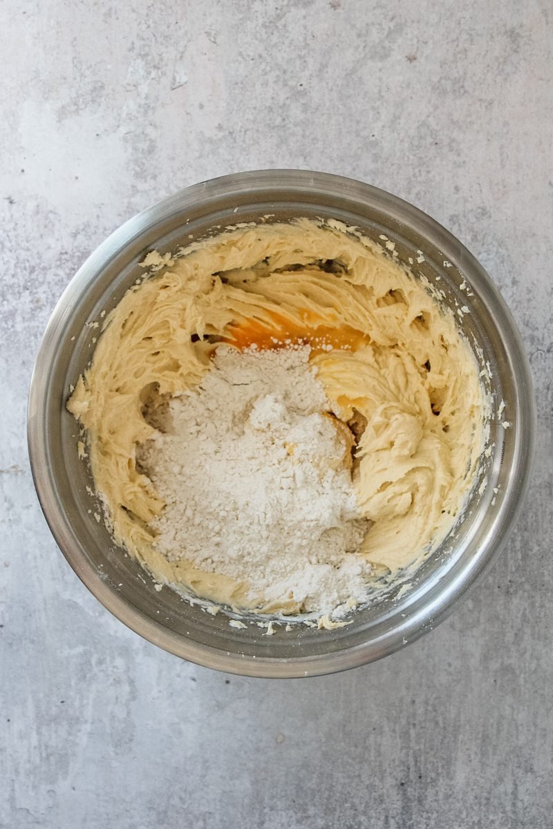 Flour and eggs sit in whipped butter in a stainless steel bowl on a gray surface.