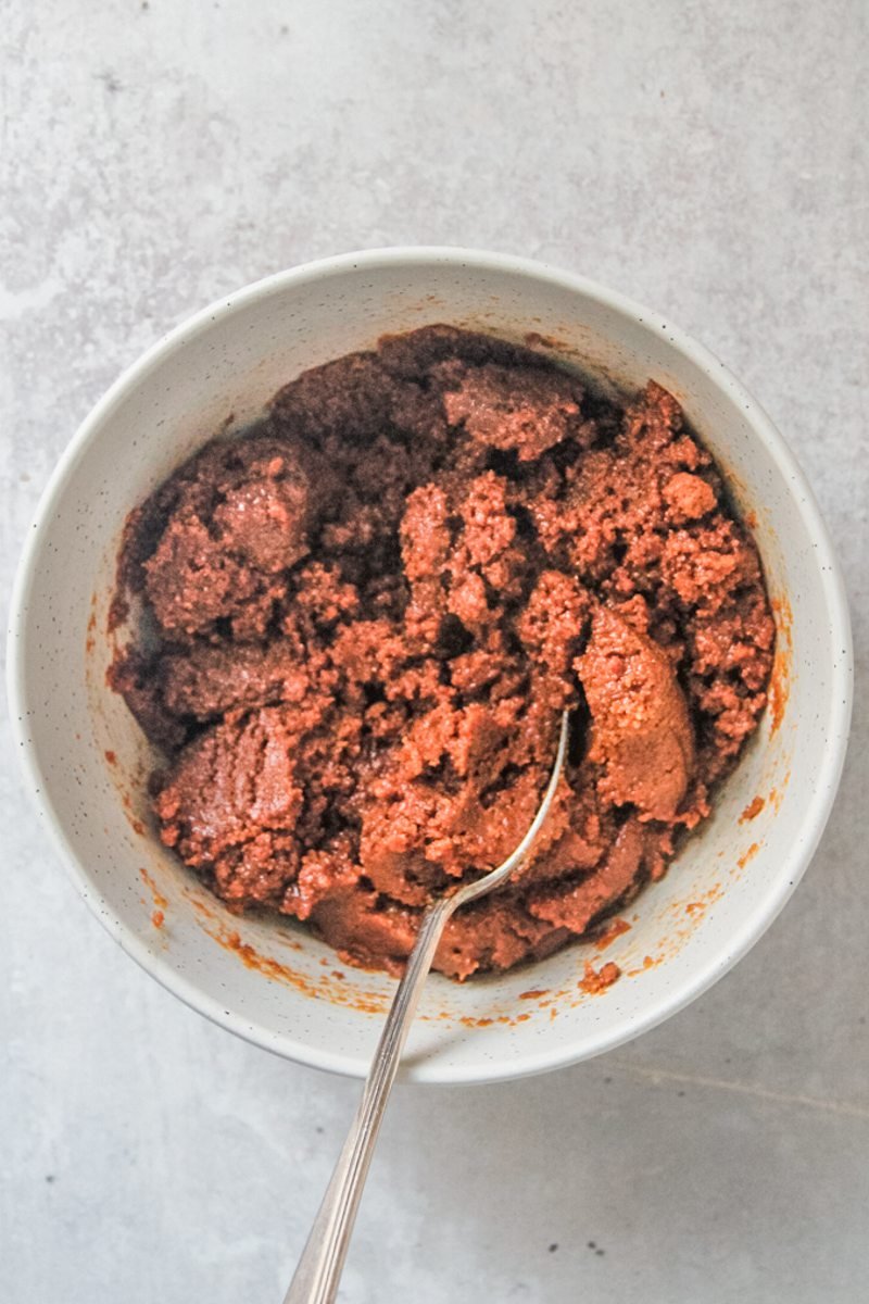 Crushed biscoff biscuits sit stirred together with melted butter in a white ceramic bowl on a gray surface.