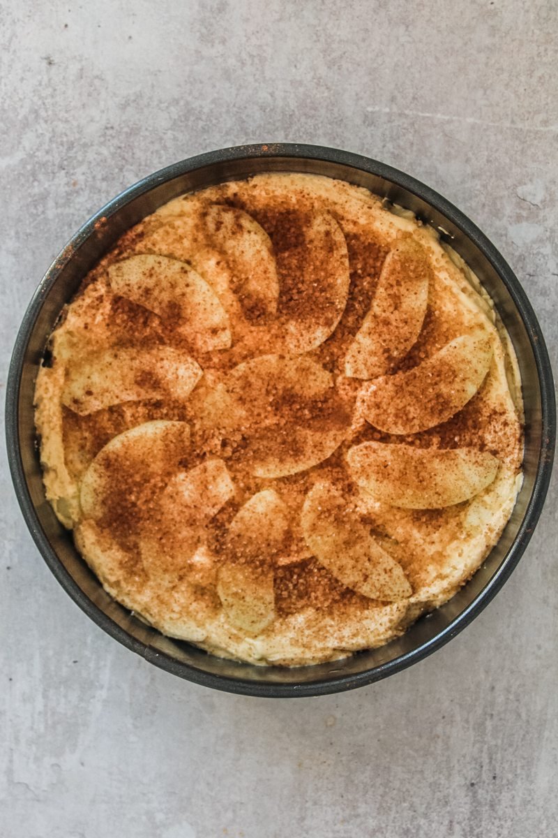 Sugar and spices sit on top of an apple cake in a round metal cake tin on a gray surface.