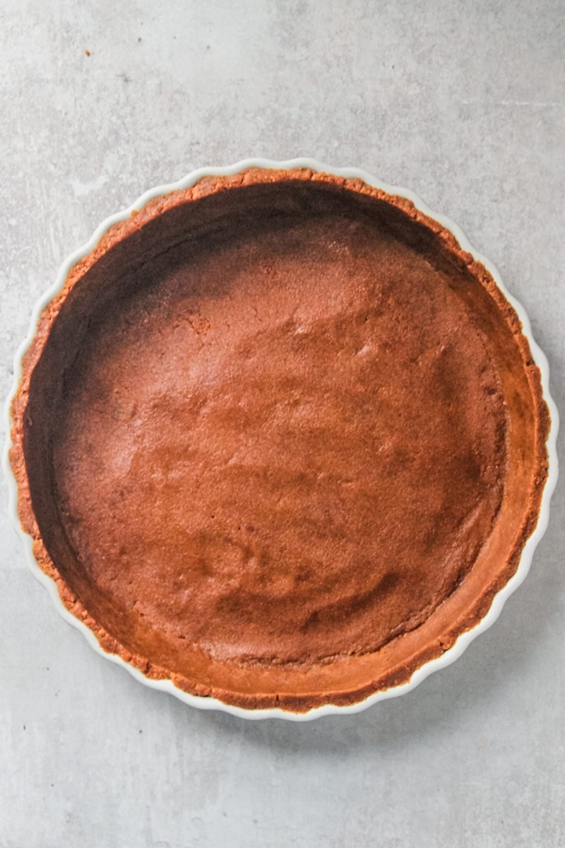 A crushed biscoff biscuit base sits pressed into a white ceramic tart dish on a gray surface.