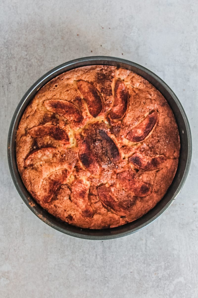 A golden apple cake with slices of apple arranged on top sit in a round metal cake tin on a gray surface.