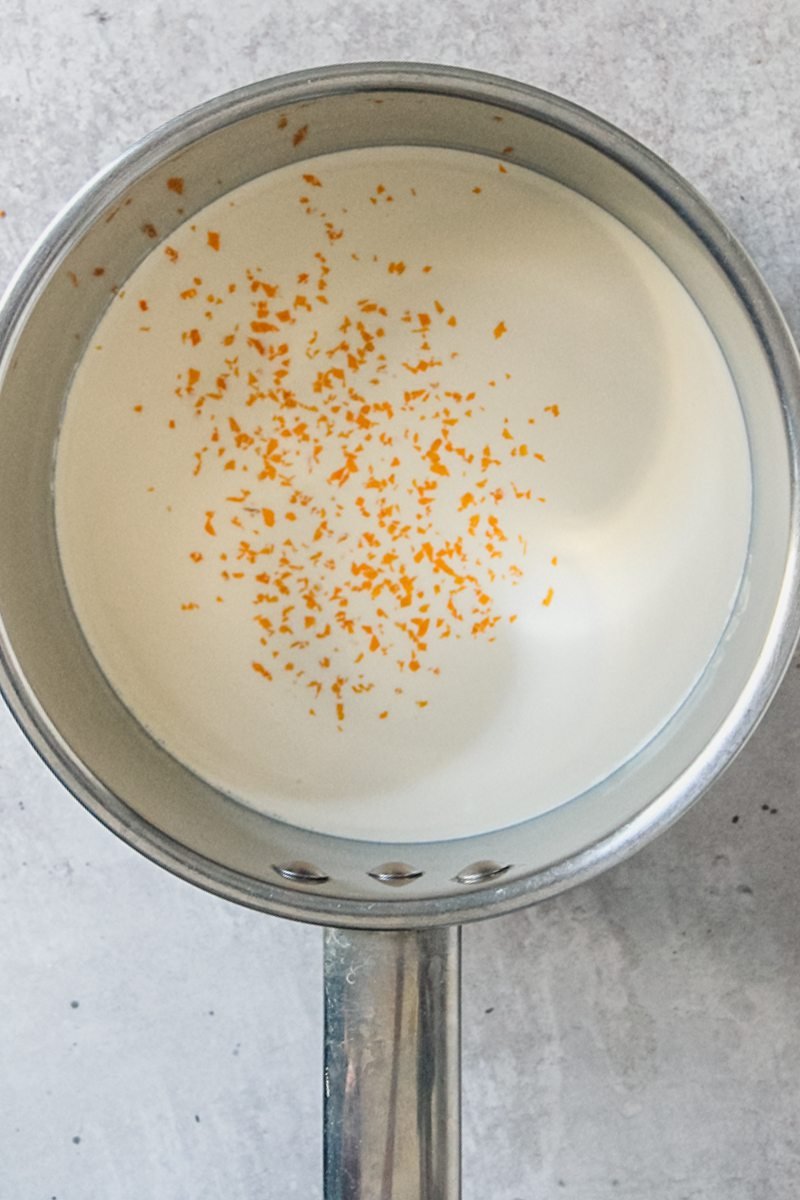 Orange zest sits in cream in a stainless steel saucepan on a gray surface.