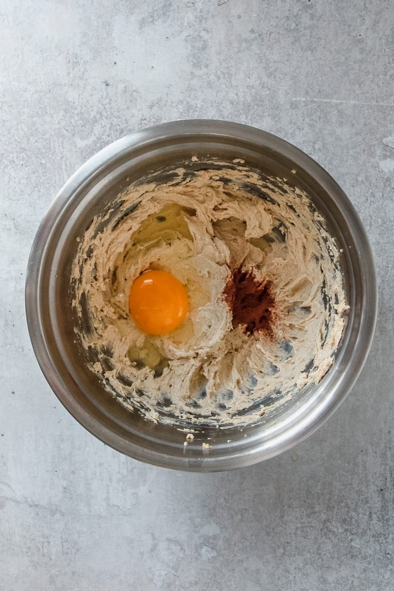 An egg and ground cinnamon sit on muffin cookie dough in a stainless steel bowl on a light grey surface.