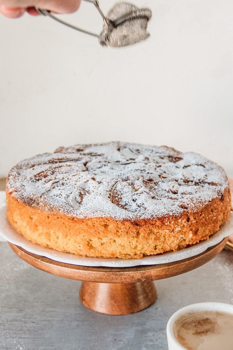 An apple cake has powdered sugar sprinkled on top as it sits on a cake stand on a gray surface.