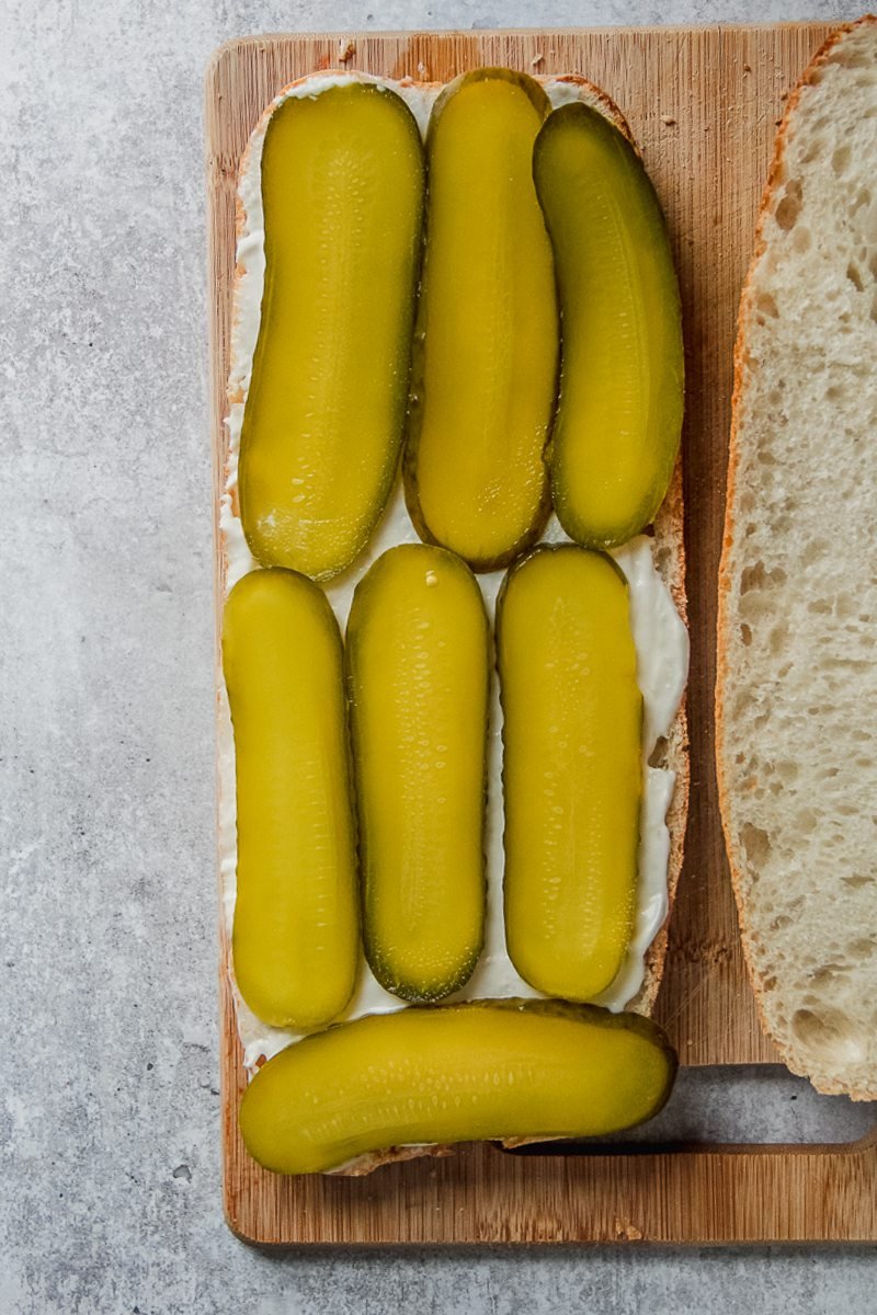 Sliced gerkins lay on top of mayonnaise on halved bread on a wooden board on a gray surface.