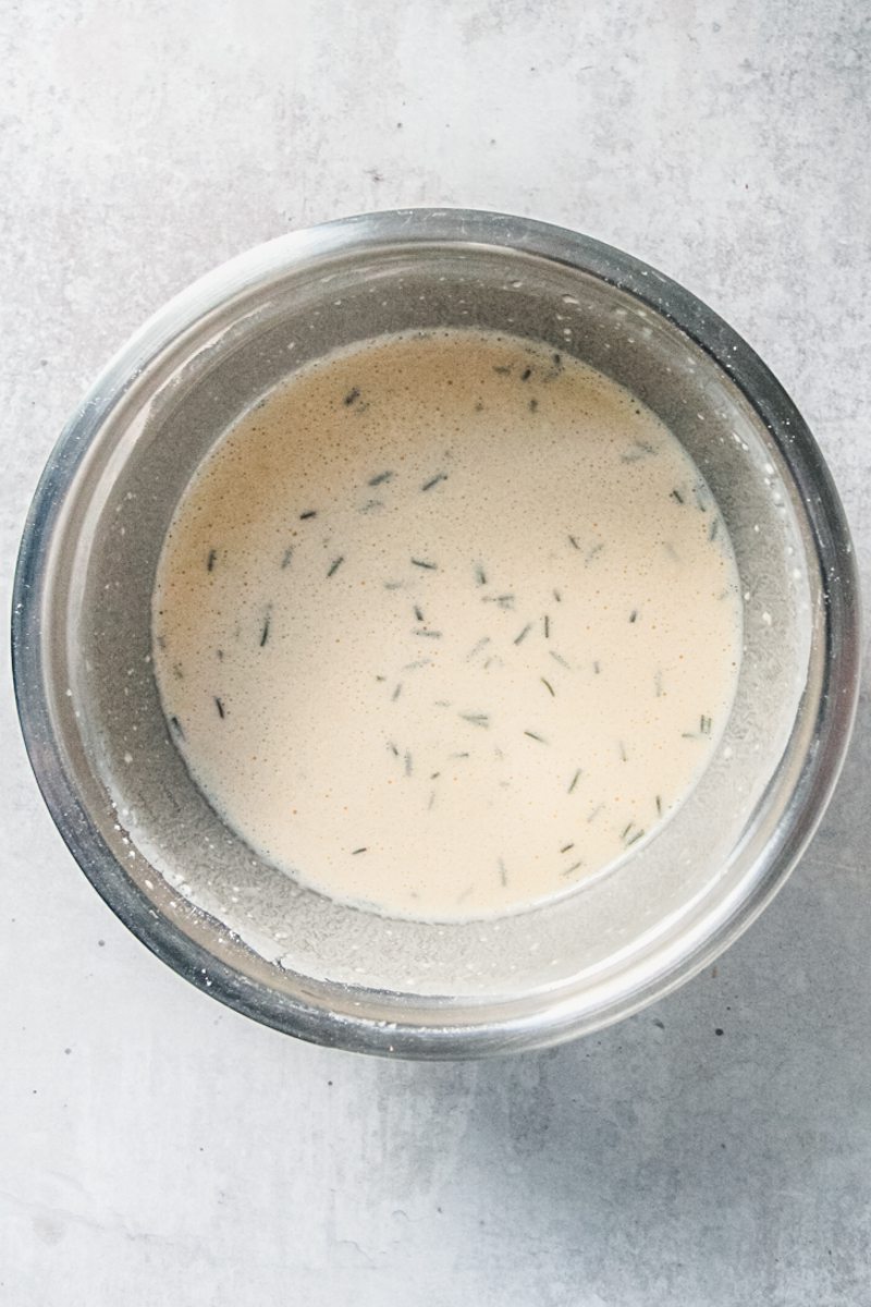 Yorkshire pudding batter with rosemary sits in a stainless steel bowl on a gray surface.