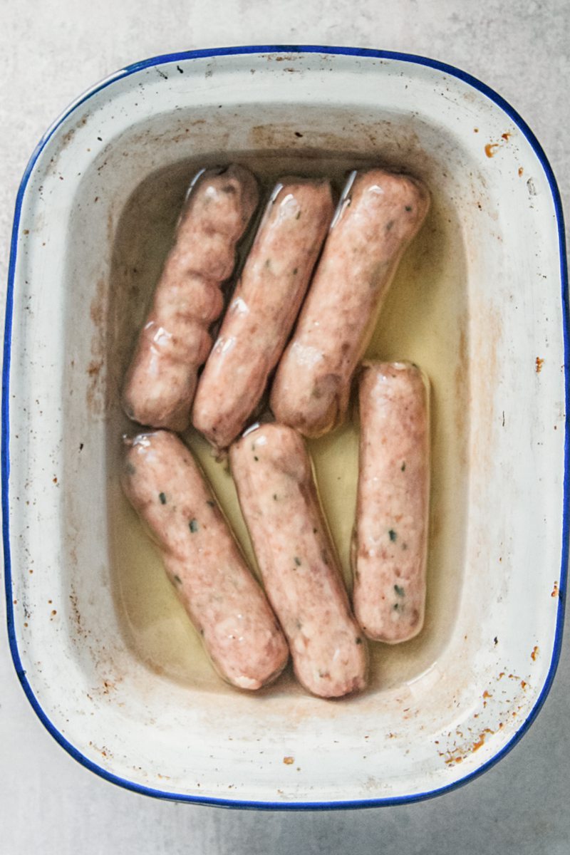 Hot oil and sausages sit in a enamel baking tin on a gray surface.