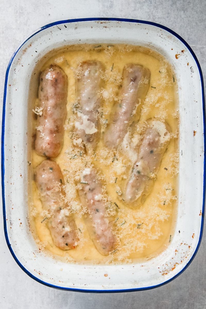 Sausages float in Yorkshire pudding batter and oil in an enamel baking tin on a gray surface.