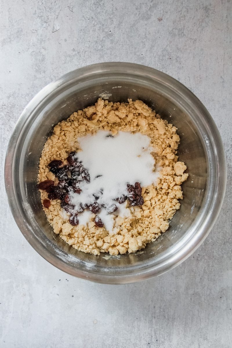 Sugar and raisins sit on top of a rough breadcrumb mixture for Welsh cakes in a stainless steel bowl on a grey surface.