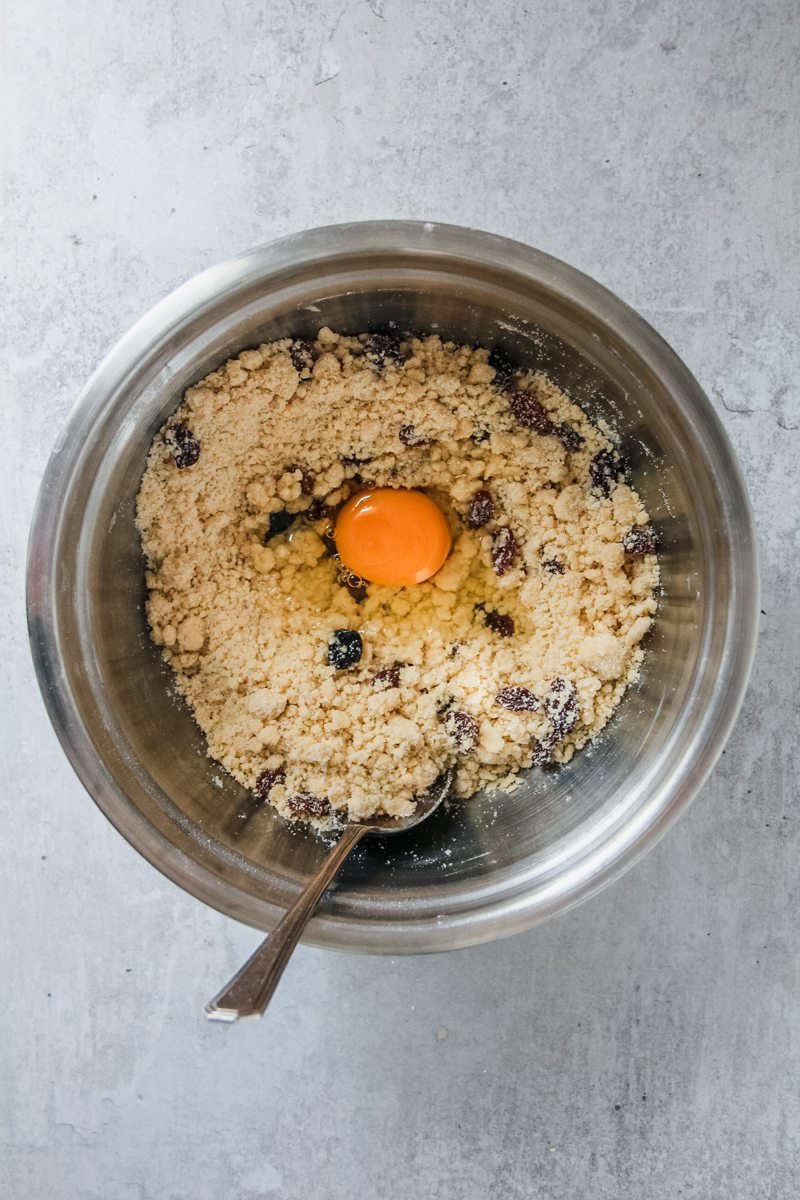 An egg sits in Welsh cake mixture in a stainless steel bowl on a grey surface.