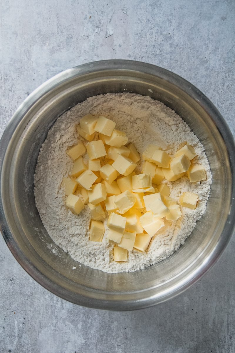 Cubes of butter sit in a flour mixture in a stainless steel bowl on a gray surface.