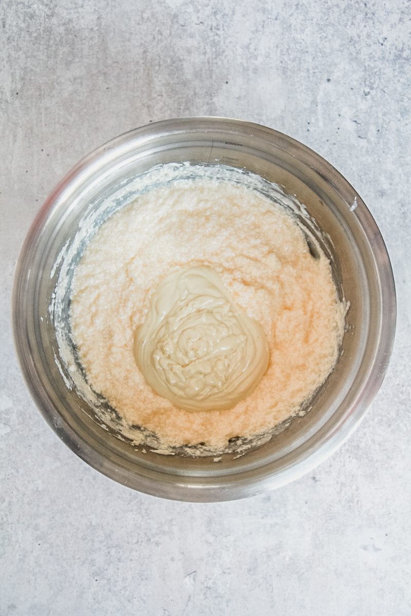 Melted chocolate sits on top of the whipped butter and sugar mixture in a stainless steel bowl on a gray surface.