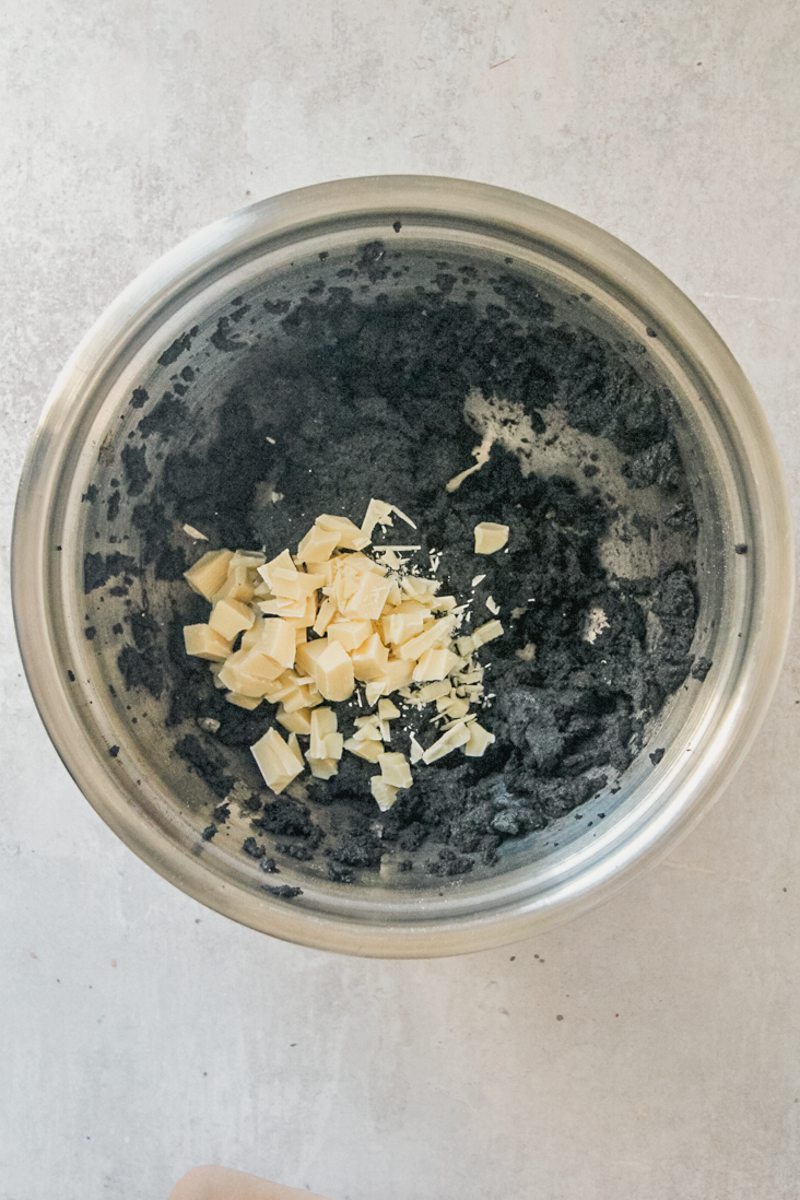 White chocolate chunks sit on top of black cocoa cookie dough in a stainless steel bowl on a gray surface.