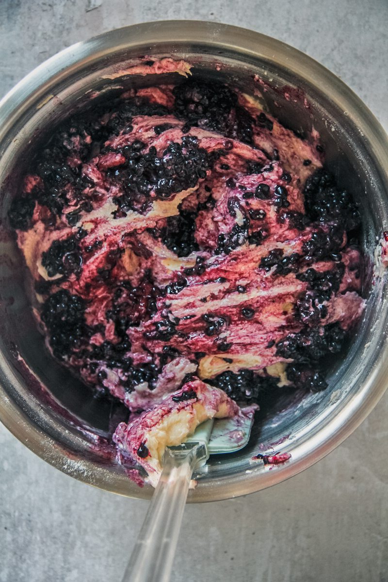 Blackberries are stirred through a white chocolate cake batter in a stainless steel bowl on a gray surface.