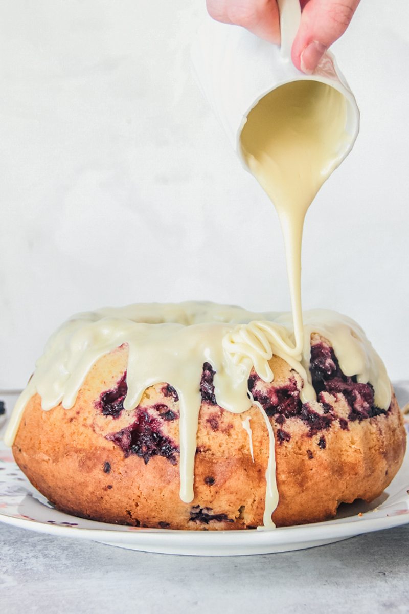 A white chocolate glaze is poured over a white chocolate blackberry bundt cake on a floral ceramic plate on a gray surface.