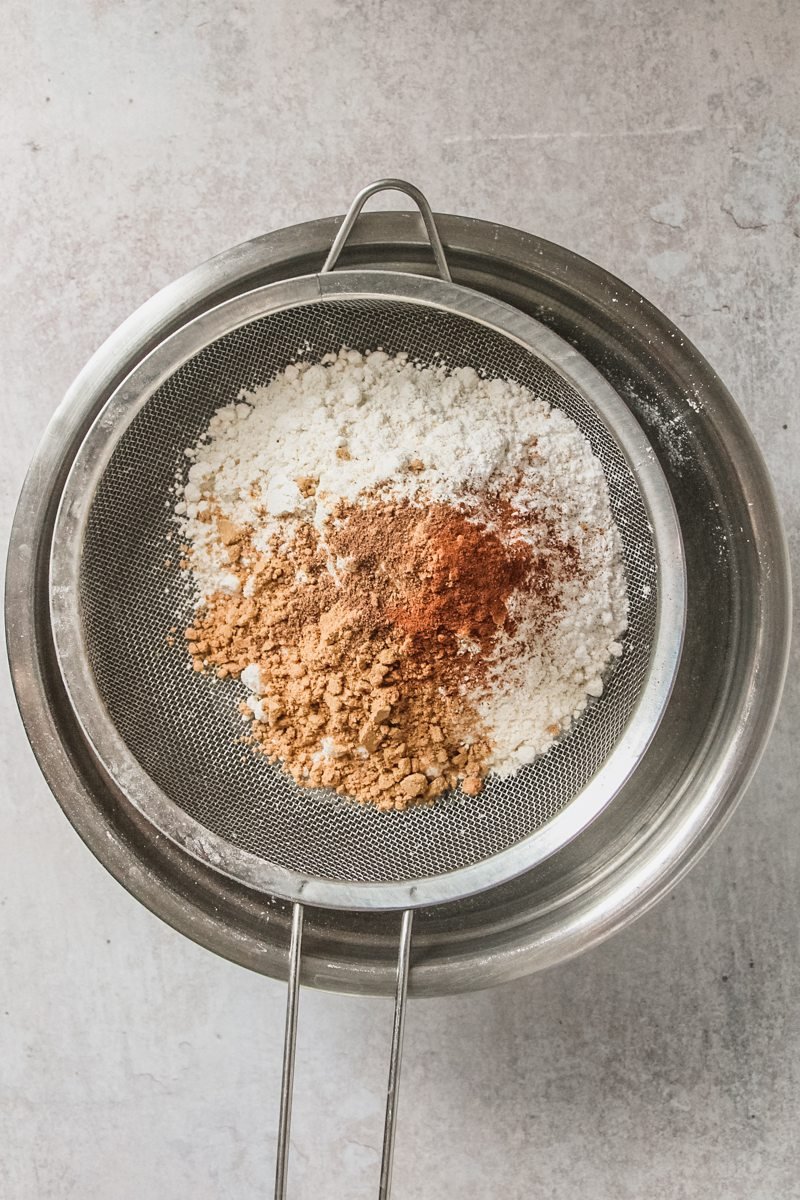 Flour and spices sit in a sift above a stainless steel bowl on a gray surface.