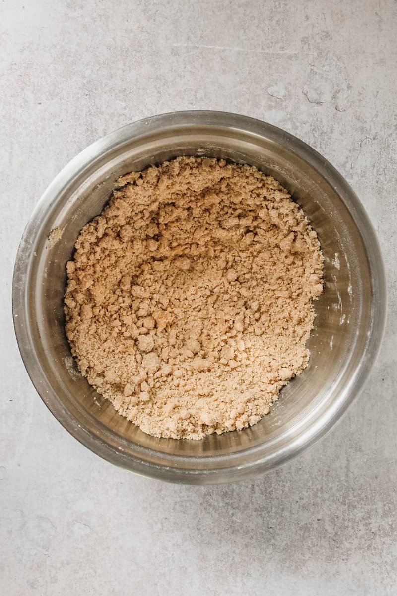 A rough breadcrumb Ginger biscuit mixture sits in a stainless steel bowl on a gray surface.