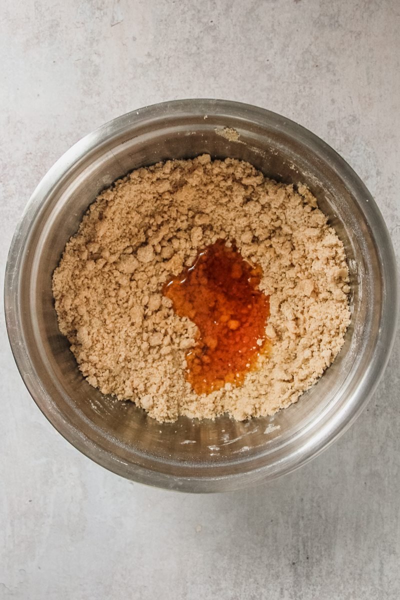 Golden syrup sits inside a rough breadcrumb ginger biscuit mixture on a gray surface.