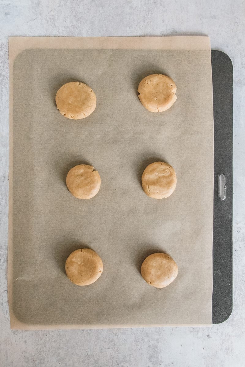 Ginger biscuit dough sits slightly flattened on a parchment paper lined baking tray on a gray surface.