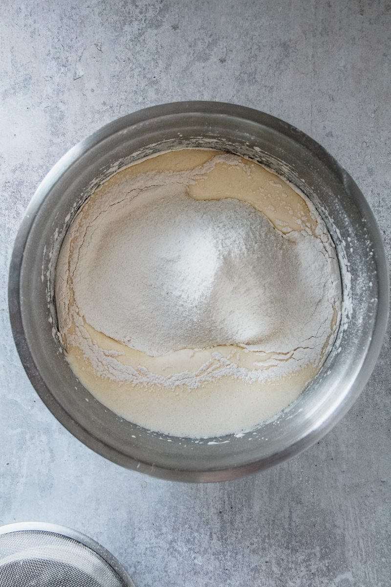 Sifted flour sits on top of a fluffy batter in a stainless steel bowl on a light gray surface.