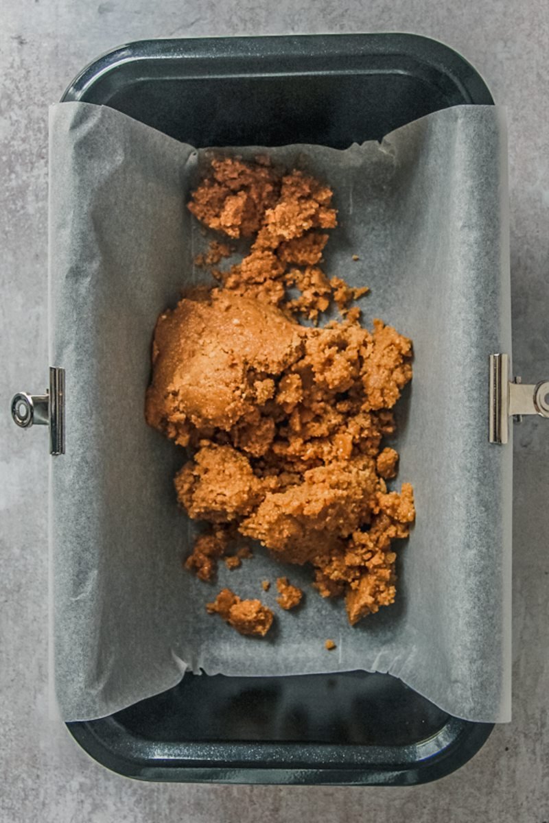 A crushed biscuit base sits in a lined metal loaf tin on a gray surface, yet to be pushed evenly into the tin.