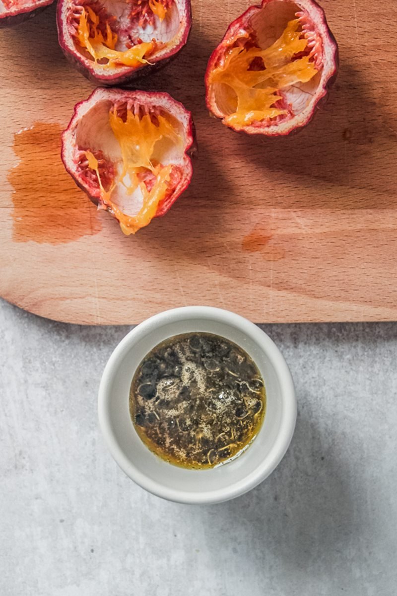 Passionfruit syrup sits in a small ceramic cup next to a wooden board and empty passionfruit.