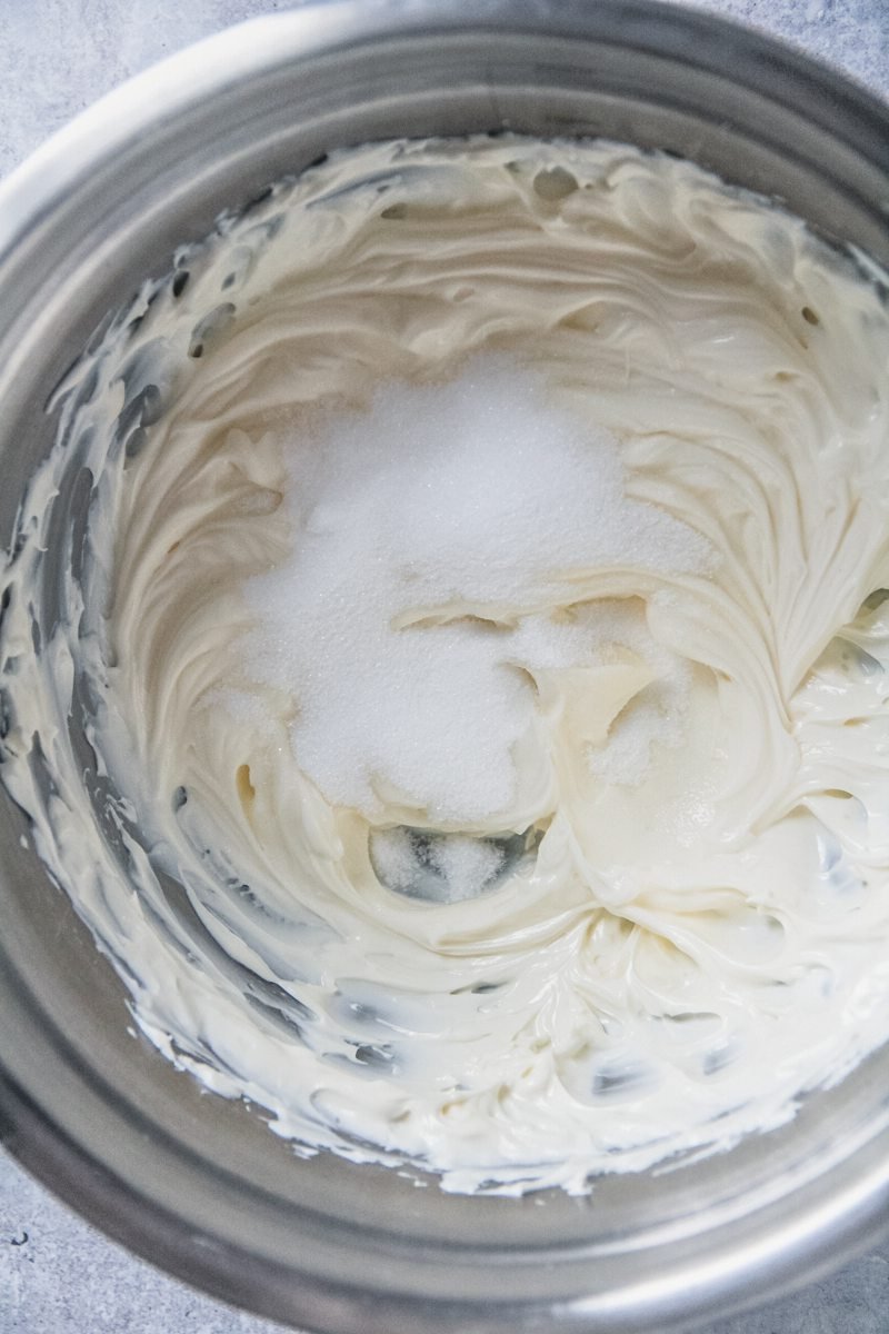 Sugar sits on top of whipped cream cheese in a stainless steel bowl on a gray surface.