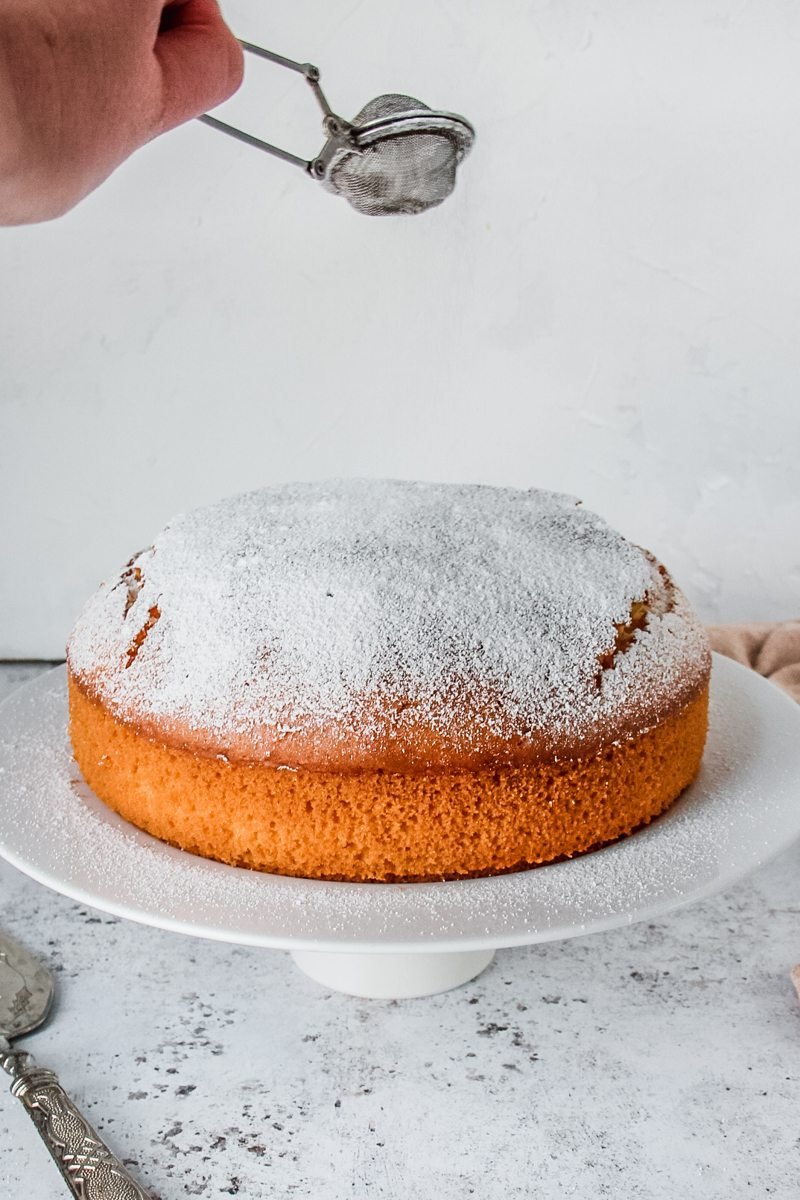 Powdered sugar is sprinkled over the top of the golden baked cake standing on a cake stand on a light gray surface.