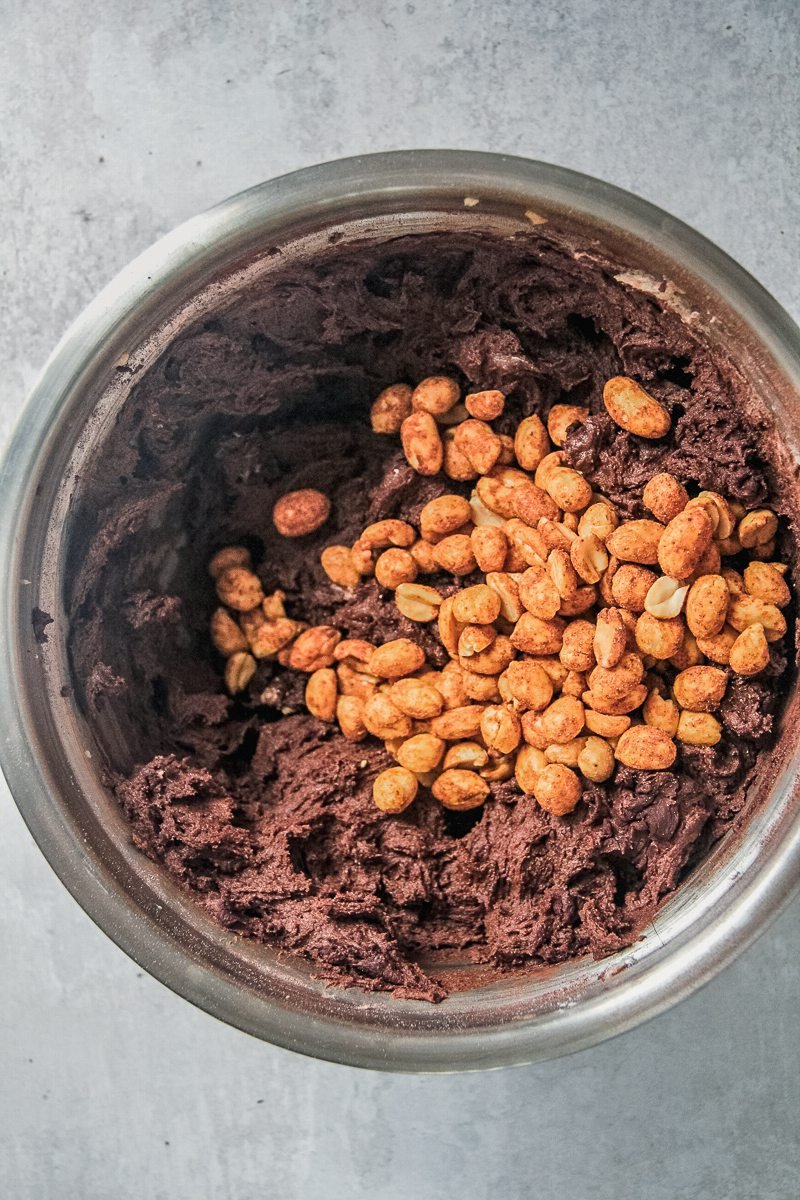 Roasted peanuts sit on top of cocoa cookie dough in a stainless steel bowl on a gray surface.