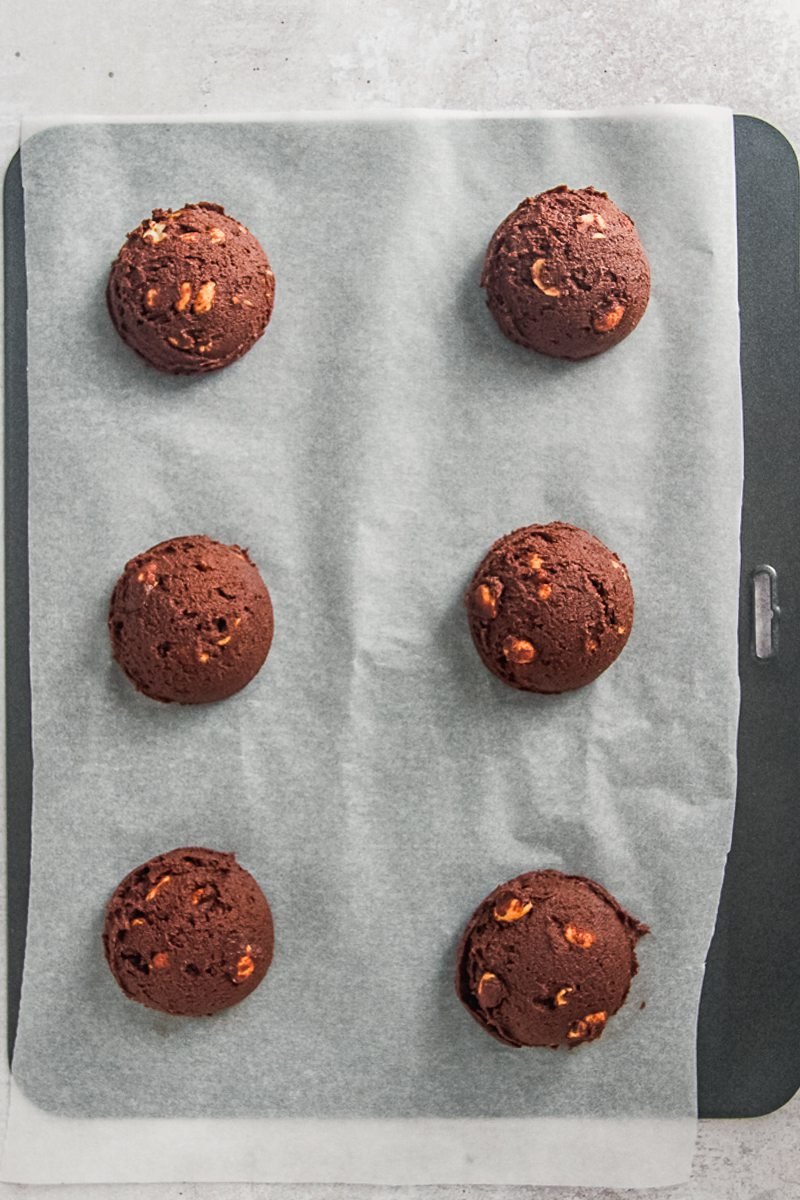 Scoops of cookie dough sit on a lined baking tray on a gray surface.