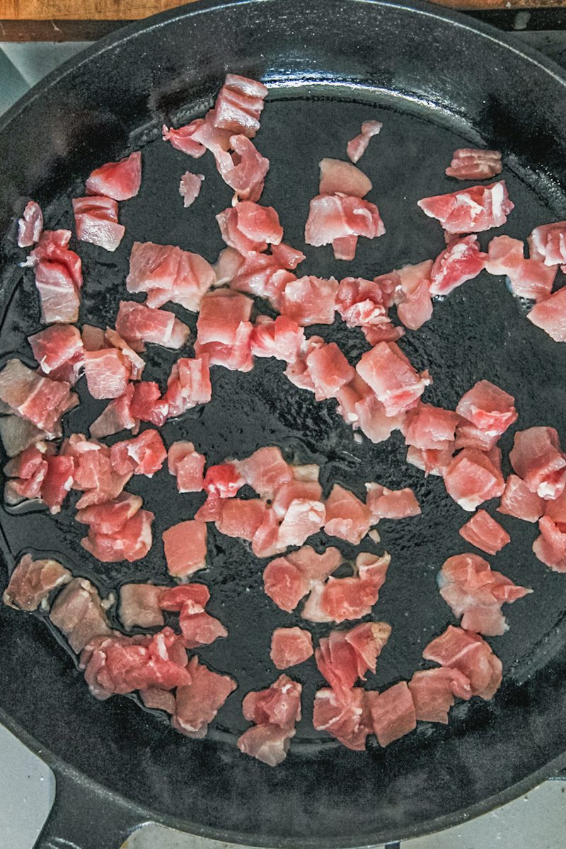 Cubes of bacon cook in a cast iron skillet on a cooker.