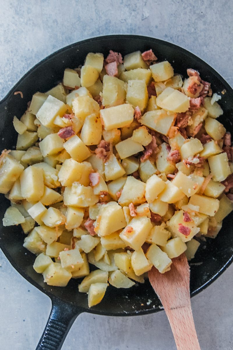 Cubes of potatoes are stirred with bacon and onion in a cast iron skillet on a gray surface.