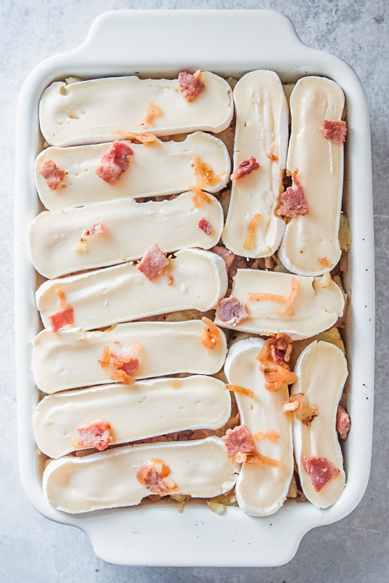 Unbaked Tartiflette sits in a rectangle white ceramic baking dish on a gray surface.