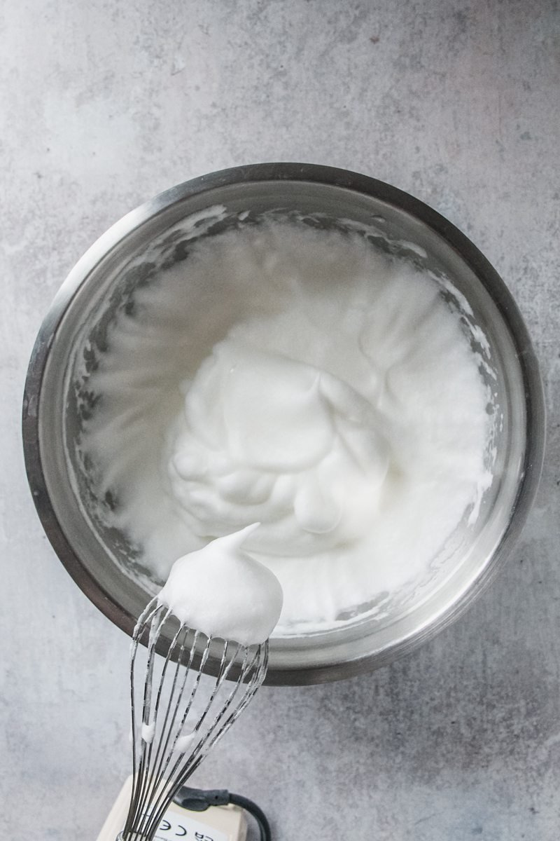 Whipped egg whites sit in a stainless steel bowl on a gray surface.