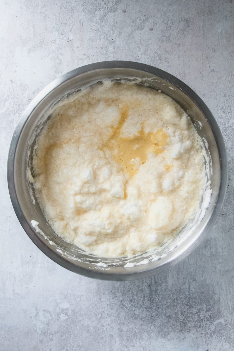 Whipped egg yolks are added to whipped egg whites and rice pudding for a cake batter in a stainless steel bowl on a light gray surface.