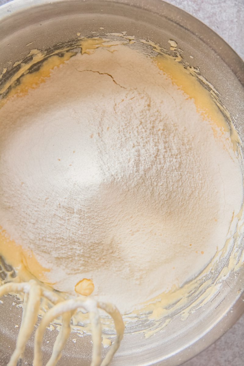 Sifted dry ingredients sit on top of a whipped cake mixture in a stainless steel bowl on a gray surface.