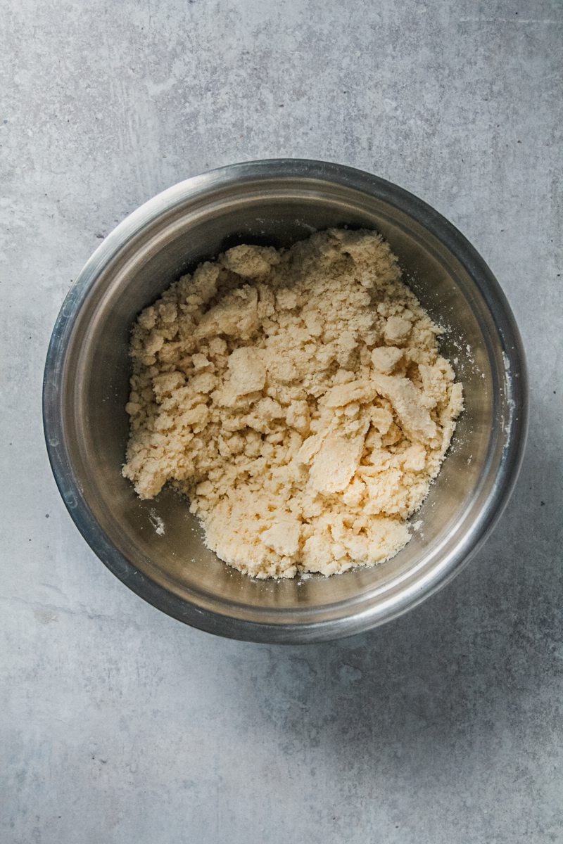 A shortbread crumble mixture sits in a stainless steel bowl on a gray surface.