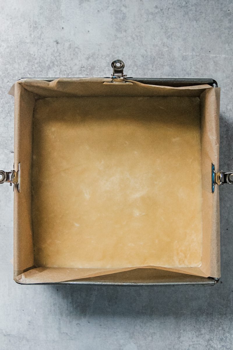 A shortbread base sits tightly packed into a lined square baking tin on a gray surface.