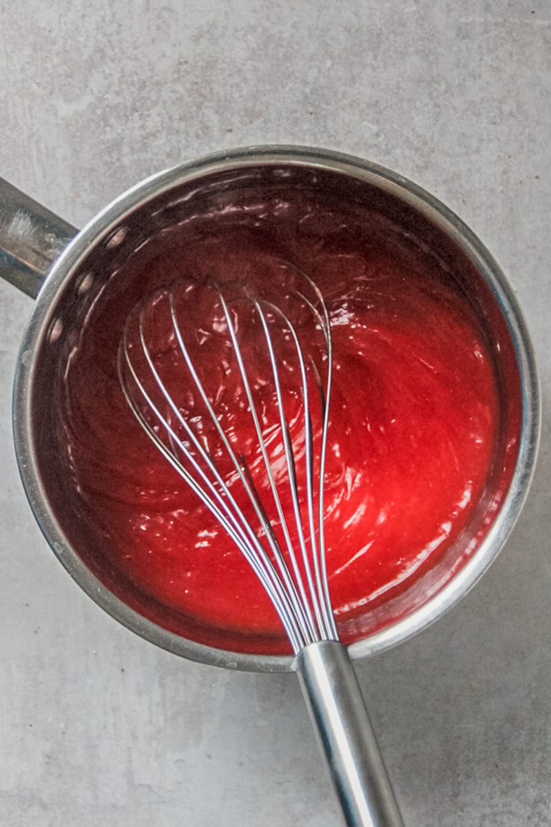 A whisked and thickened blood orange juice mixture sits in a stainless steel saucepan on a gray surface.