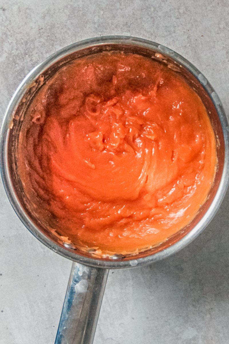 A blood orange curd sits in a stainless steel saucepan on a gray surface.