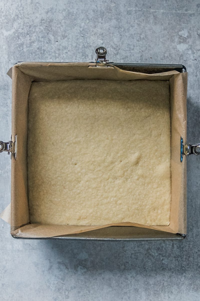 A baked shortbread base sits in a lined square metal baking tin on a gray surface.