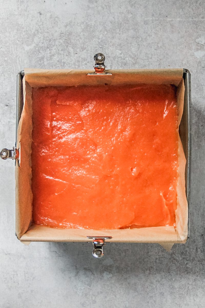 An unbaked blood orange curd sits in a lined square baking tin on a gray surface.