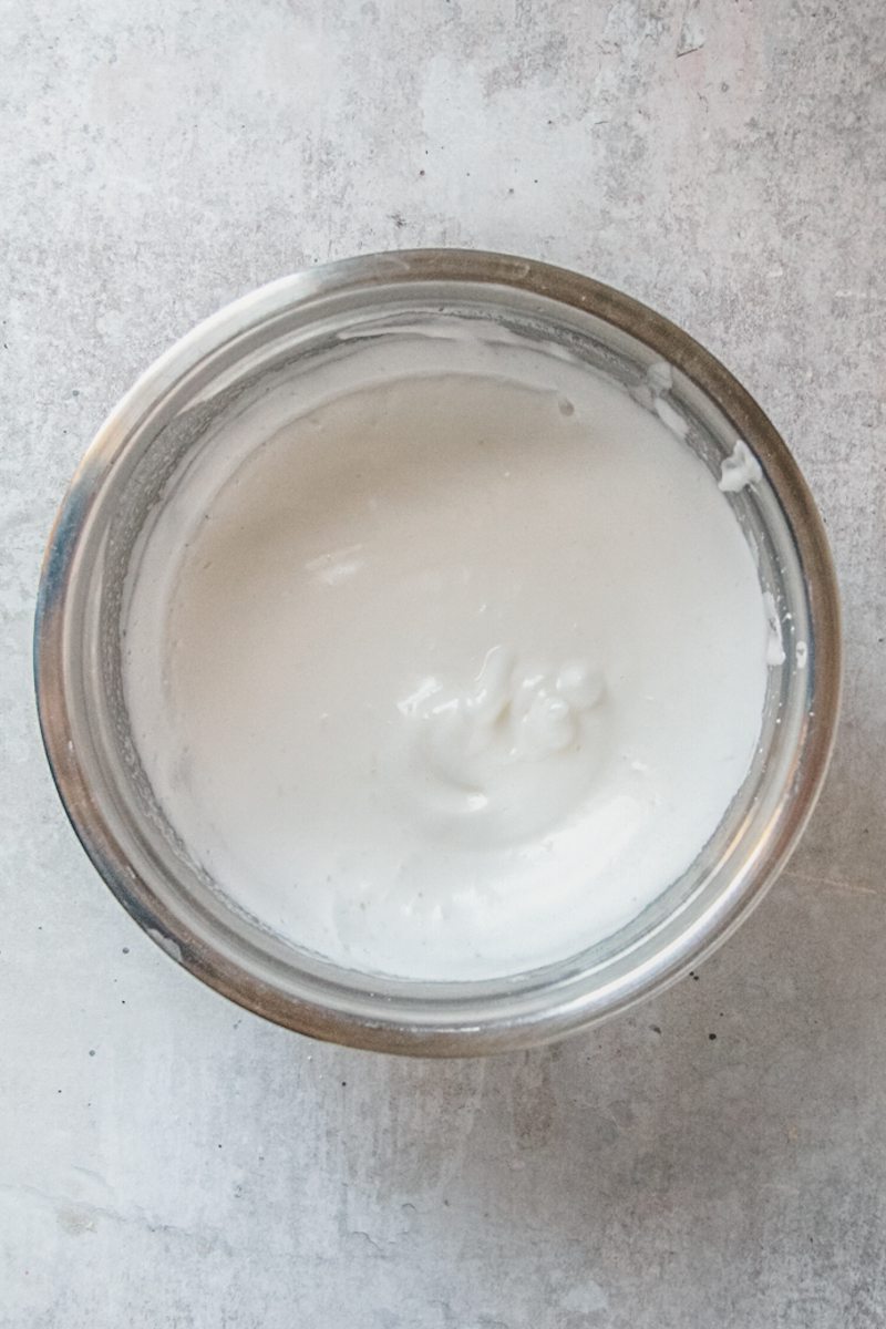 Whipped swiss meringue sits in a stainless steel bowl on a light grey background.