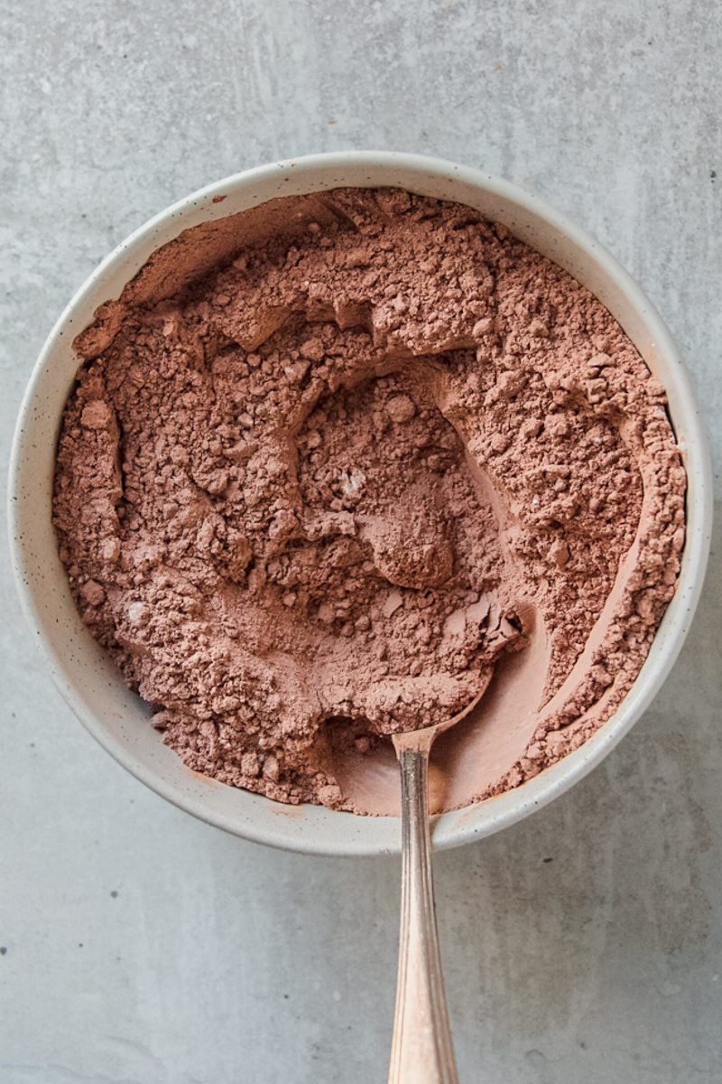 Flour and cocoa sit stirred in a ceramic bowl on a gray surface.