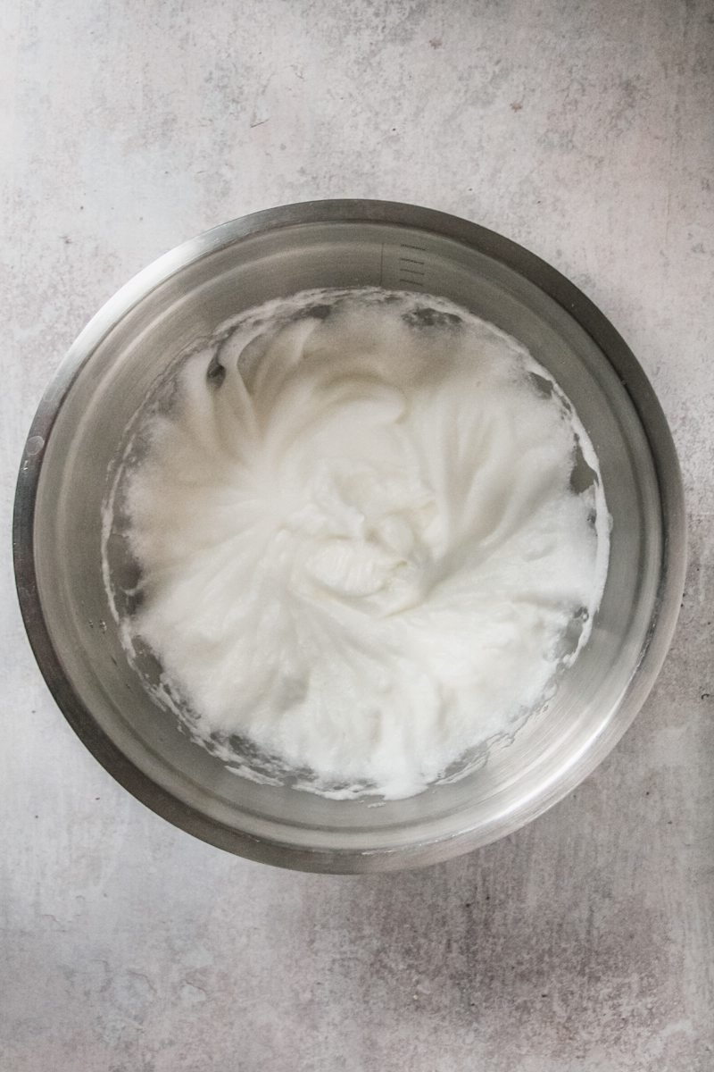 Whipped egg whites sit in a stainless steel bowl on a gray surface.