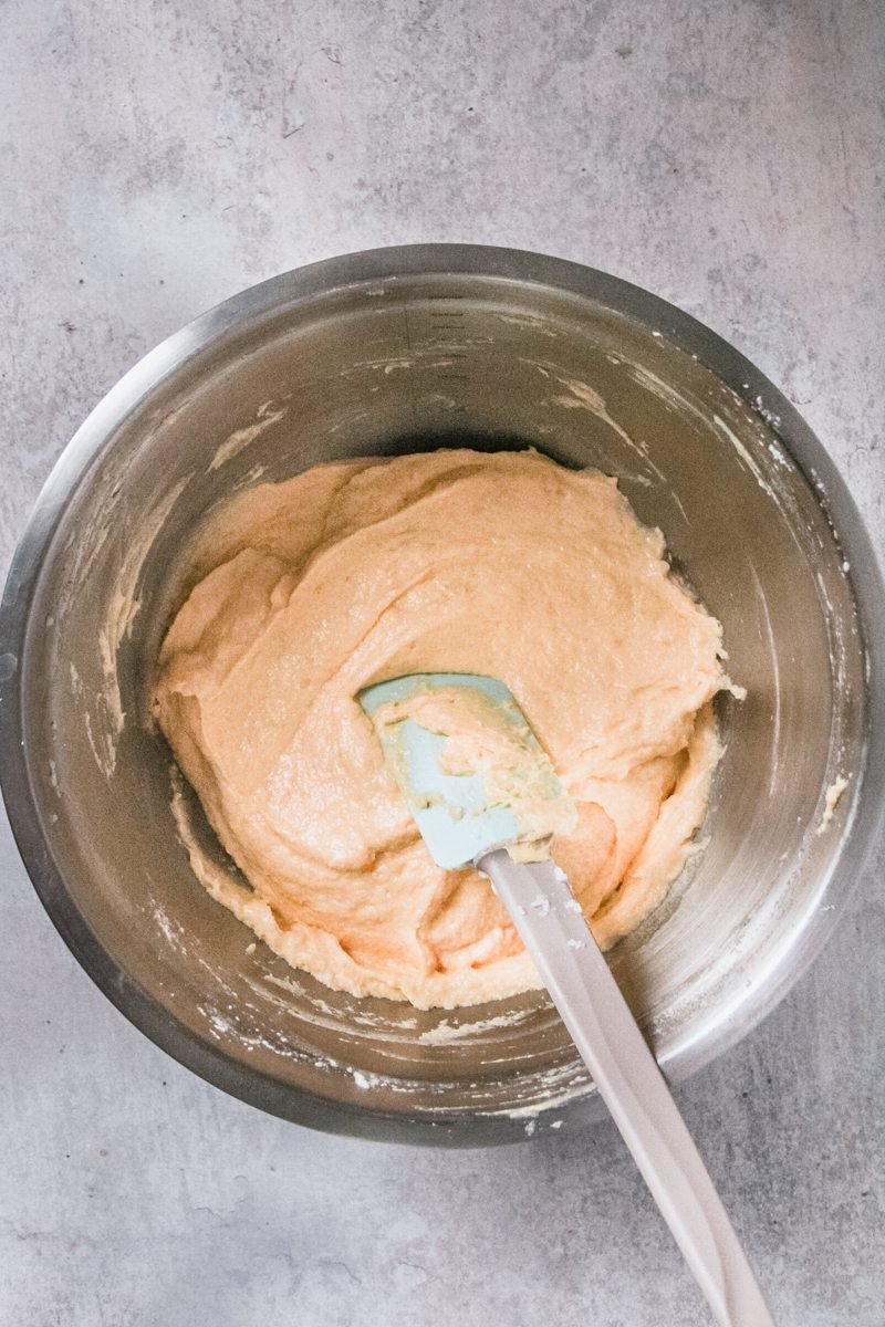 Cake batter sits in a stainless steel bowl with a spatula sitting on the batter on a gray surface.